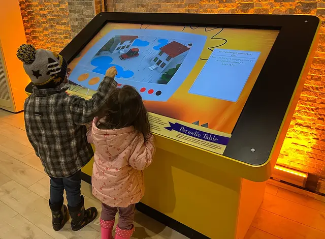 Two children using the Periodic Table touchscreen to explore a water-compound example.
